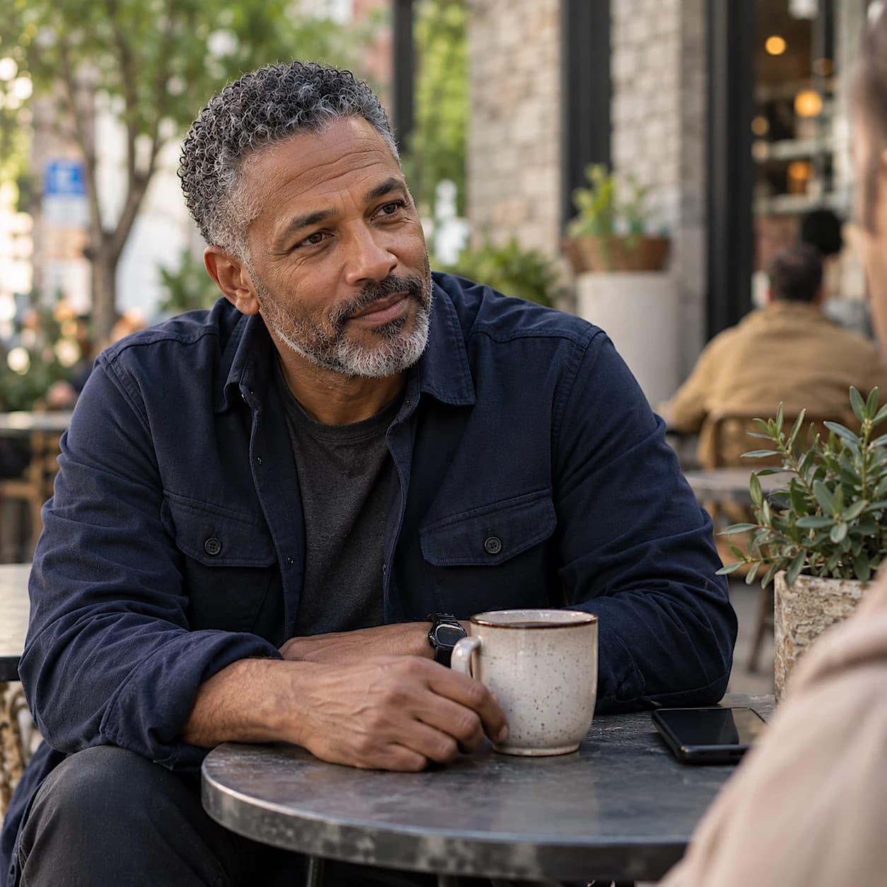 A man with short gray curly hair and a gray beard sits at an outdoor café table with a coffee mug, listening to someone across from him with a gentle smile